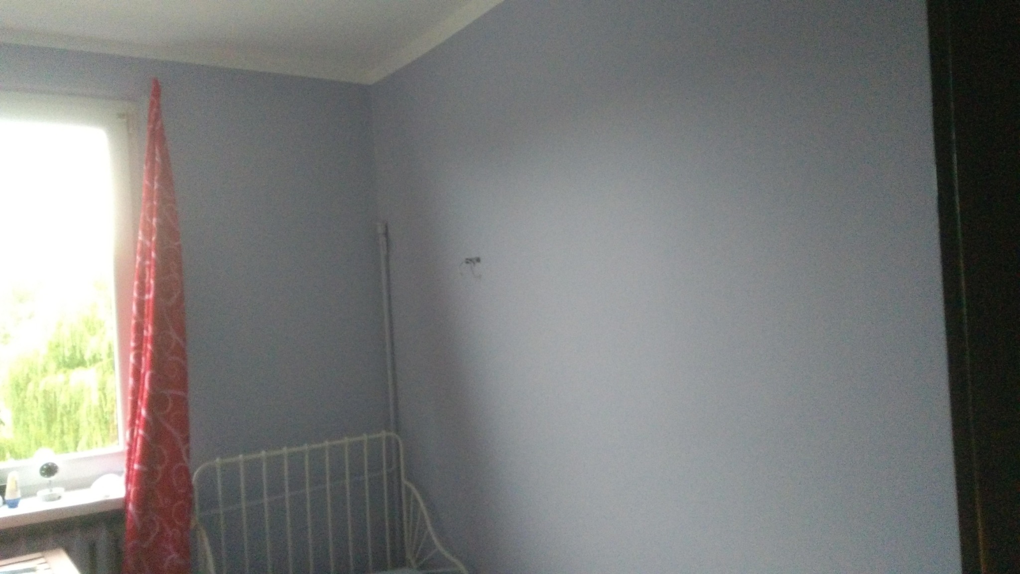 Świeżo pomalowana ściana na szaro w pokoju z widocznym fragmentem okna, zasłony w czerwone wzory i metalowego zagłówka łóżka.