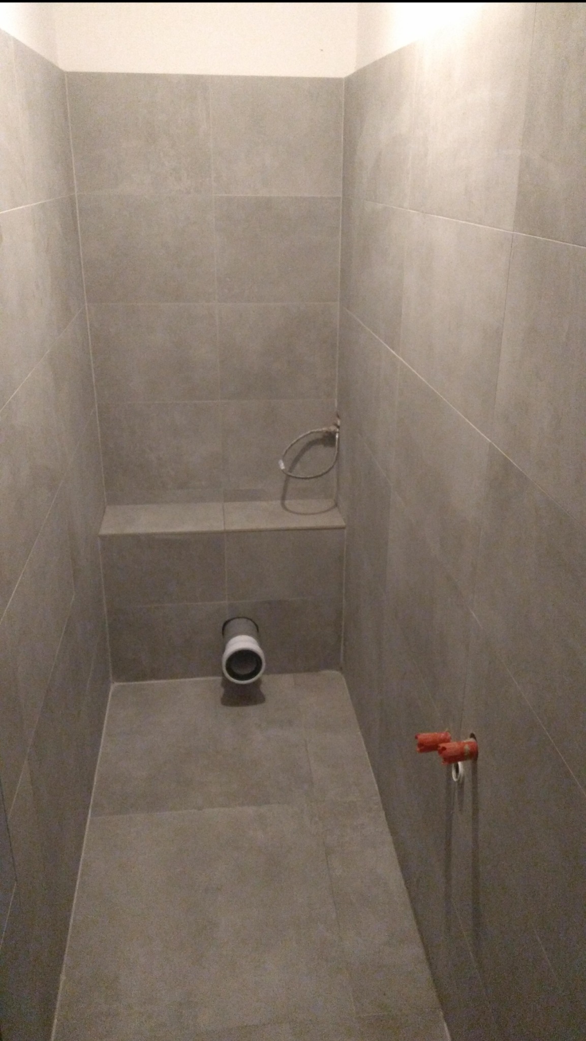 Wnętrze łazienki w trakcie remontu, widoczne szare płytki na ścianach i podłodze, wystające rury z pomarańczowymi zaślepkami oraz rura kanalizacyjna w ścianie, przygotowana do montażu toalety.