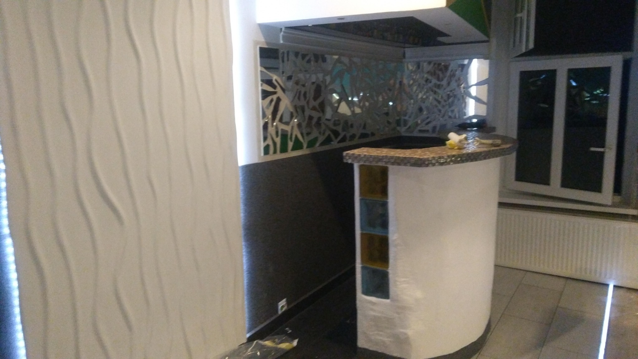 Nowoczesna ściana z trójwymiarową fakturą obok baru z mozaikową górą i kolorowymi wstawkami, widok na okno i dekoracyjną ściankę ażurową w tle.