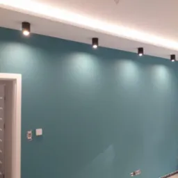 Ściana pomalowana na turkusowo z rzędem czarnych, punktowych lamp sufitowych, widoczne gniazdka elektryczne i fragment białej ściany oraz drzwi do innego pomieszczenia.