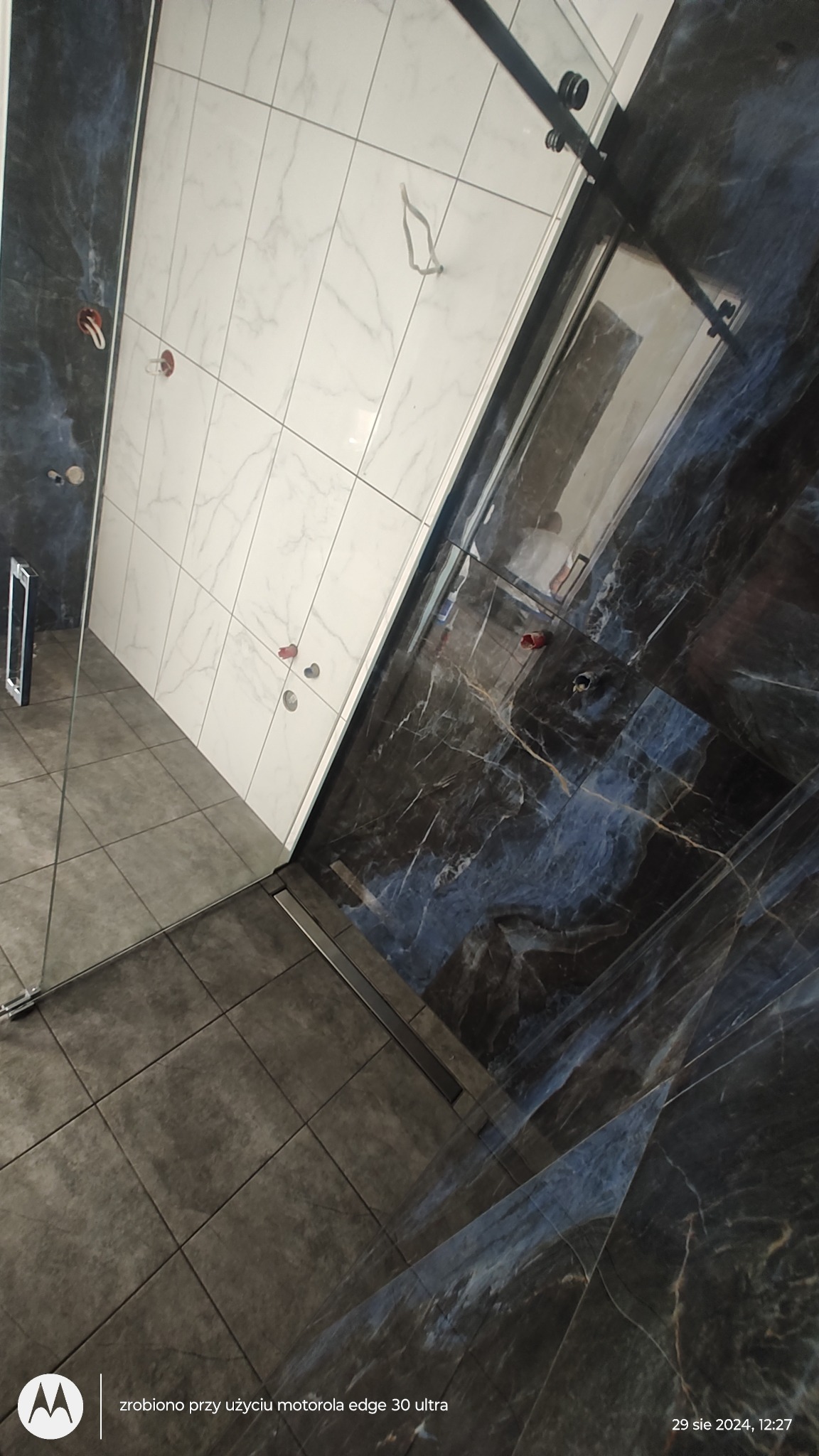 Nowoczesna kabina prysznicowa z czarnymi marmurowymi płytkami i białymi płytkami imitującymi marmur na ścianach, widoczny odpływ liniowy i szklane drzwi.