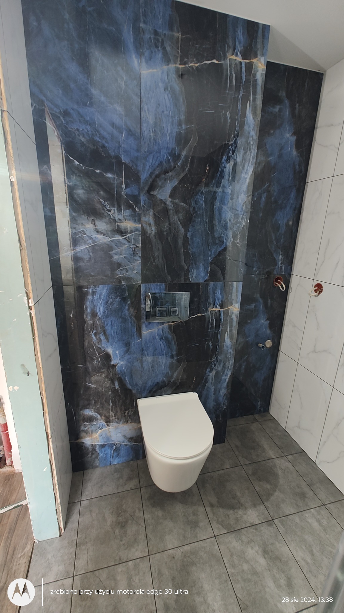 Wnętrze łazienki w trakcie prac wykończeniowych, widoczna toaleta wisząca na tle ściany wyłożonej płytkami imitującymi marmur w odcieniach granatu i szarości, obok fragment ściany z białych płytek...