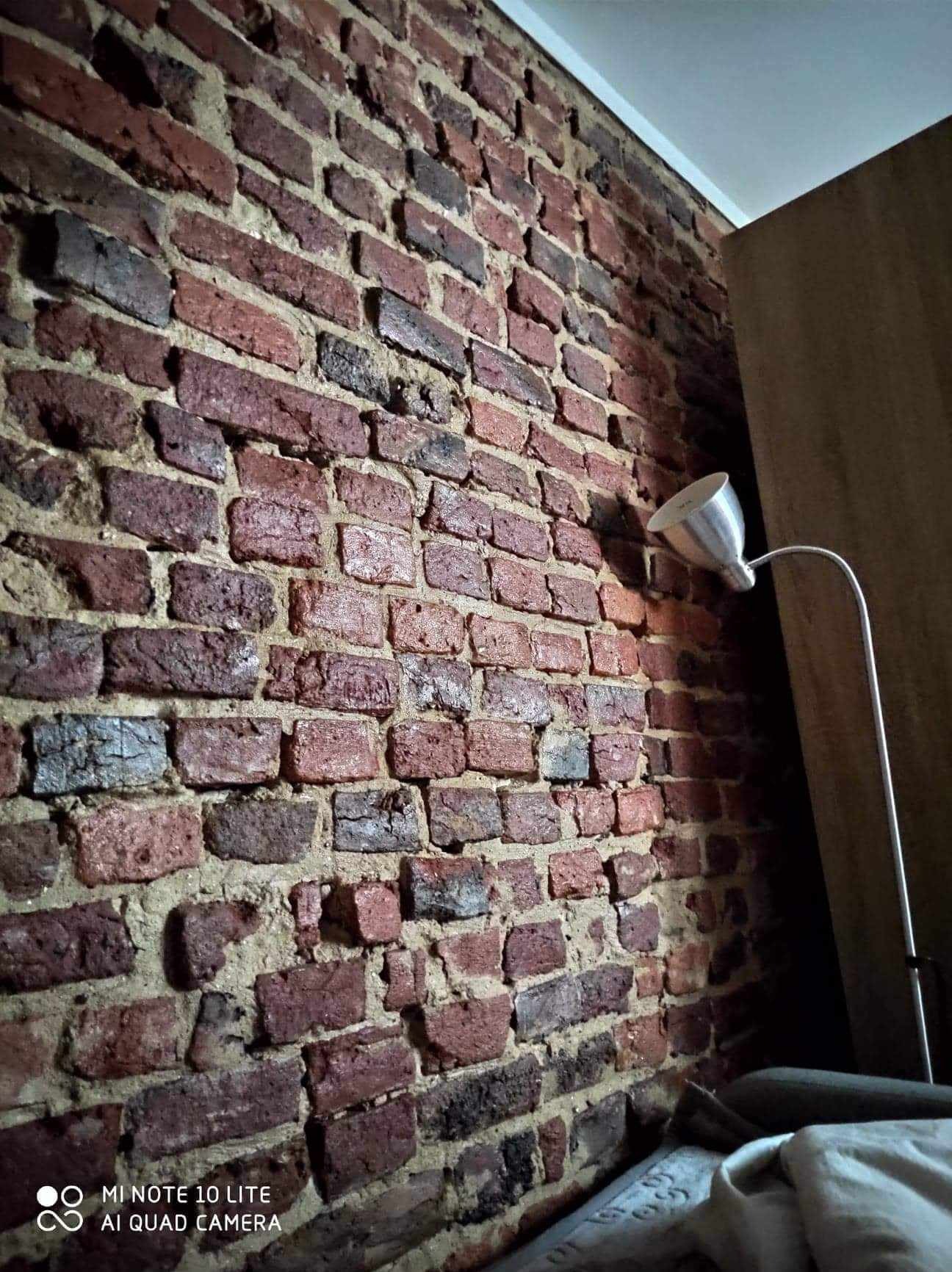 Ceglana ściana wewnętrzna z widoczną strukturą i nierównościami, oświetlona metalową lampą stojącą z białym kloszem, widok z dołu.