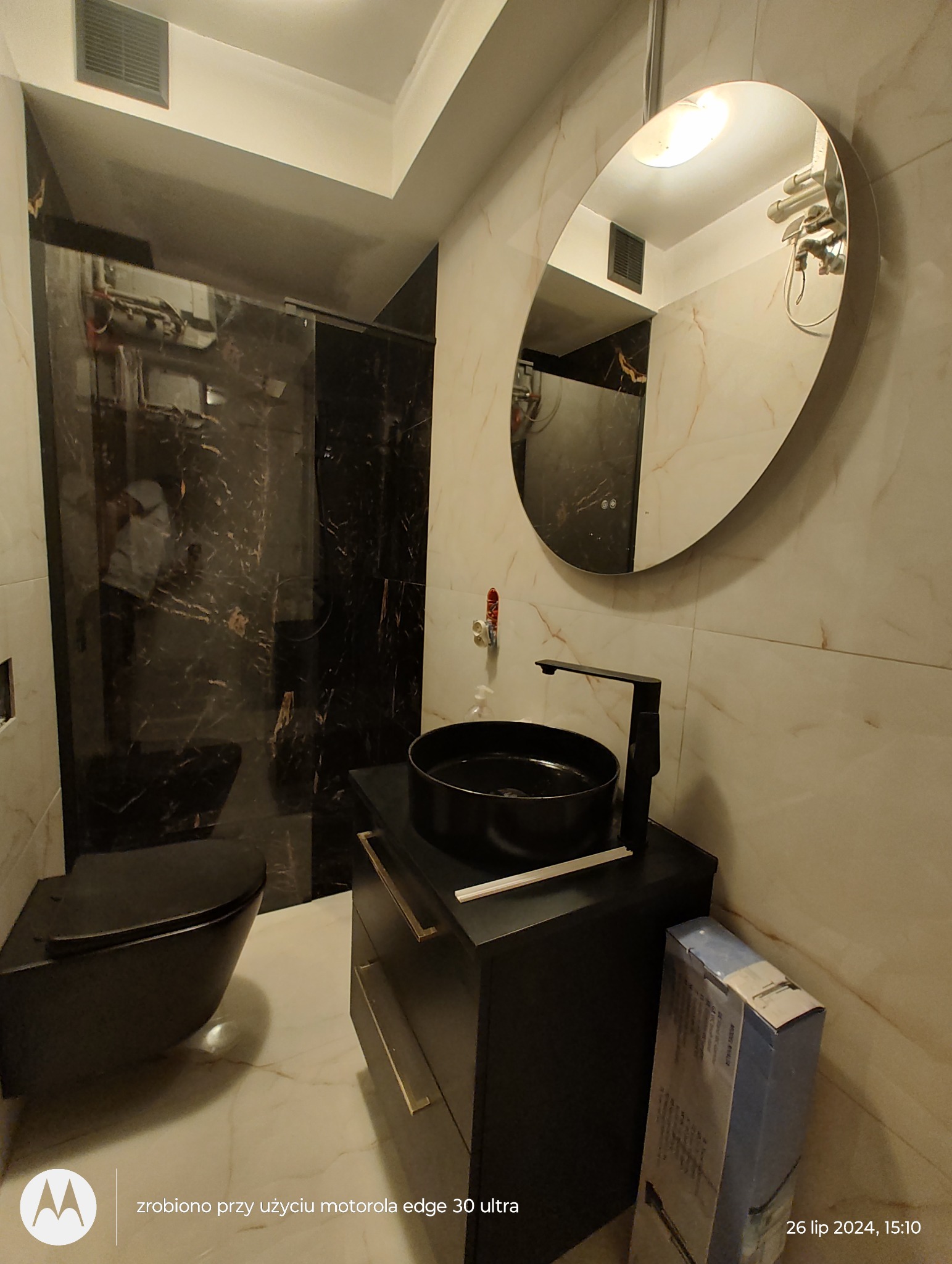 Nowoczesna łazienka w czerni i bieli z owalnym lustrem, czarną umywalką nablatową i wiszącą toaletą, marmurowe płytki na ścianach i podłodze, pudełko z akcesoriami do łazienki obok szafki...