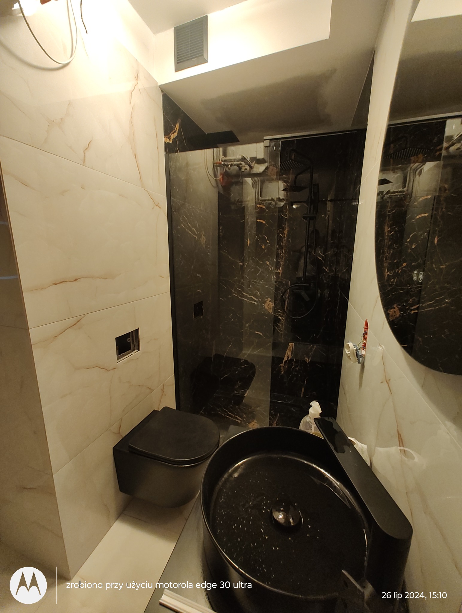 Nowoczesna łazienka z czarną armaturą, marmurowe płytki na ścianach, kabina prysznicowa typu walk-in z czarnymi profilami, wisząca toaleta i umywalka w kolorze czarnym, widoczne niedokończone...