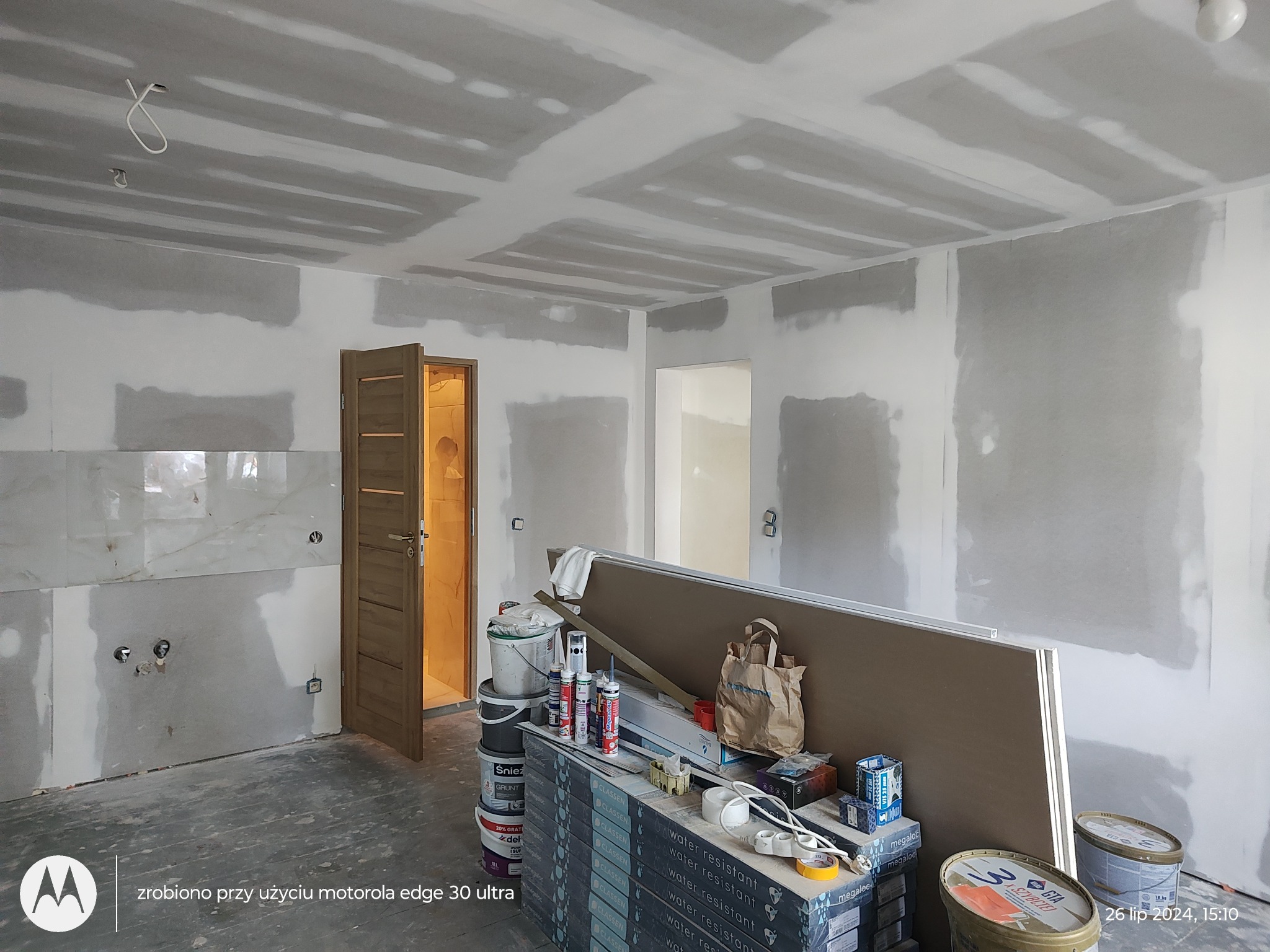 Wnętrze w trakcie szpachlowania ścian i sufitu w Pabianicach, widoczne płyty gipsowo-kartonowe, wiaderka z masą szpachlową, puszki z pianką montażową oraz otwarte drzwi do innego pomieszczenia.
