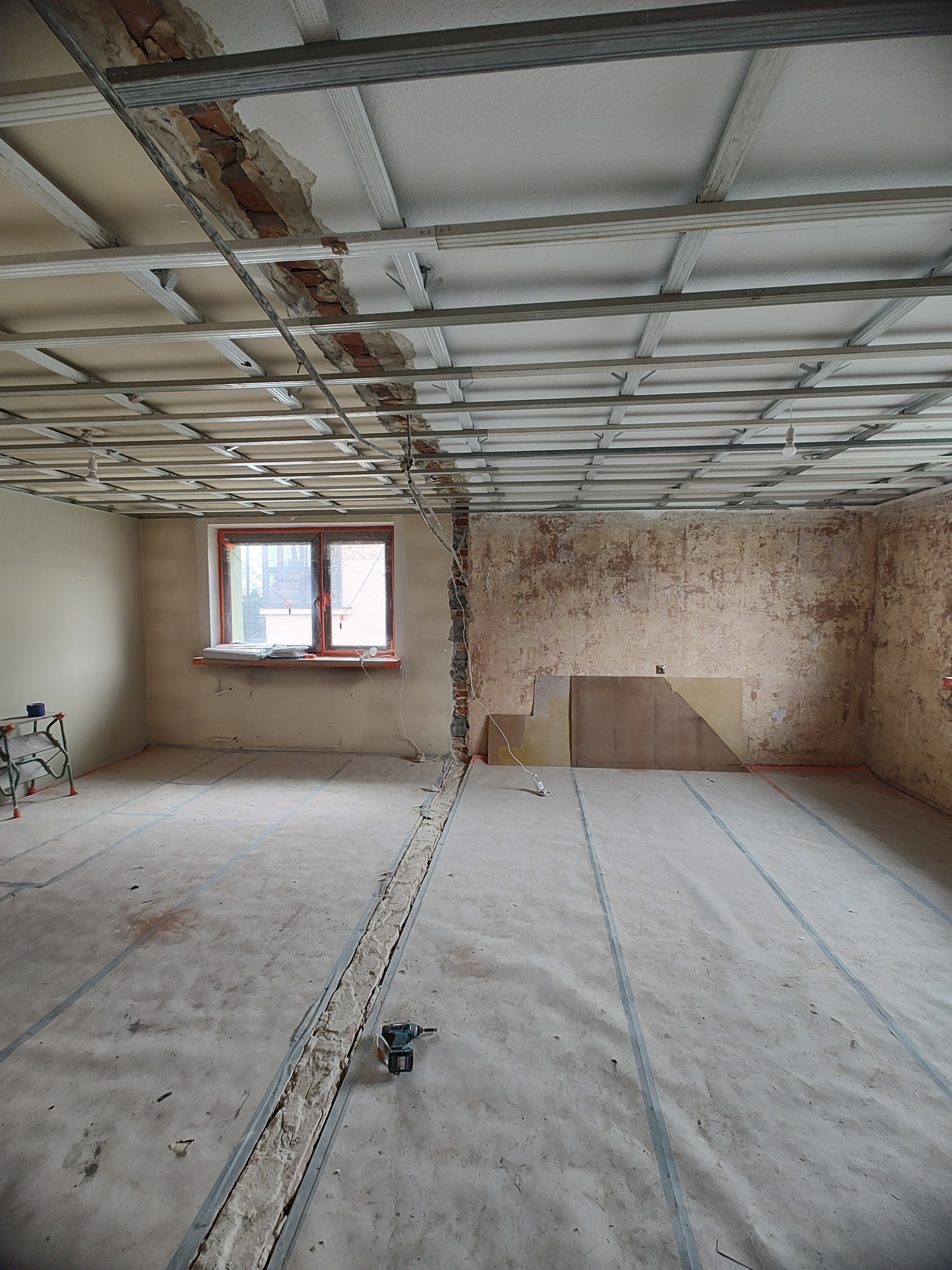 Pokój w trakcie prac wykończeniowych: widoczna konstrukcja sufitu podwieszanego z odsłoniętymi fragmentami cegieł, ślady po usuniętych ścianach, przygotowane podłoże pod panele oraz narzędzia...