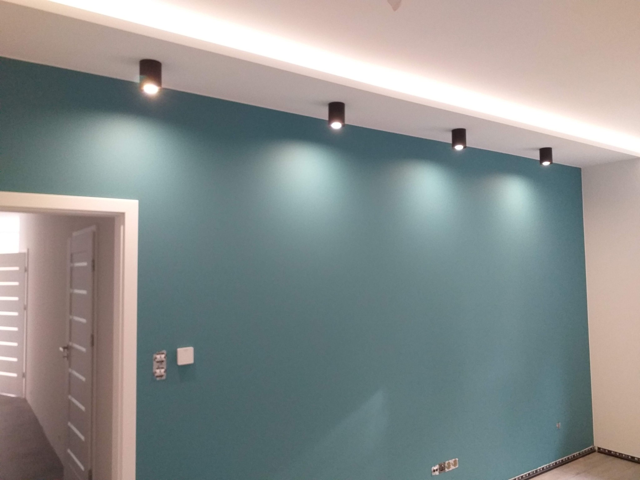Ściana pomalowana na turkusowo z rzędem czarnych, punktowych lamp sufitowych, widoczne gniazdka elektryczne i fragment białej ściany oraz drzwi do innego pomieszczenia.