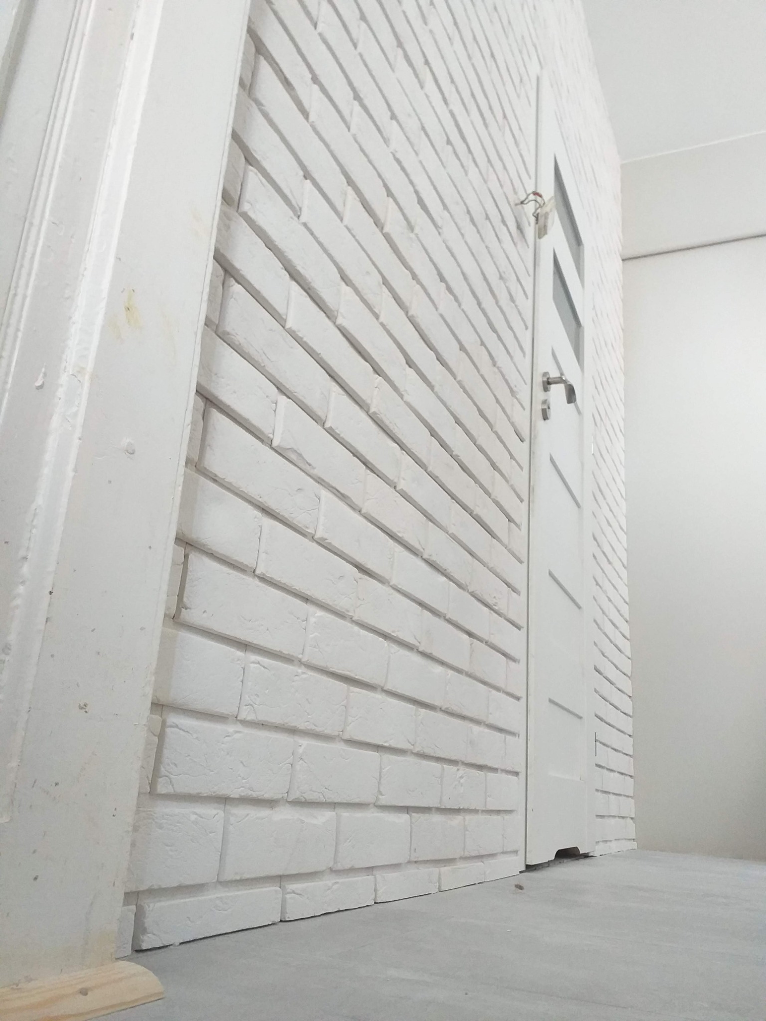 Ściana z białej cegły dekoracyjnej, białe drzwi z przeszkleniem w trakcie montażu, widoczny fragment ościeżnicy i surowa posadzka.
