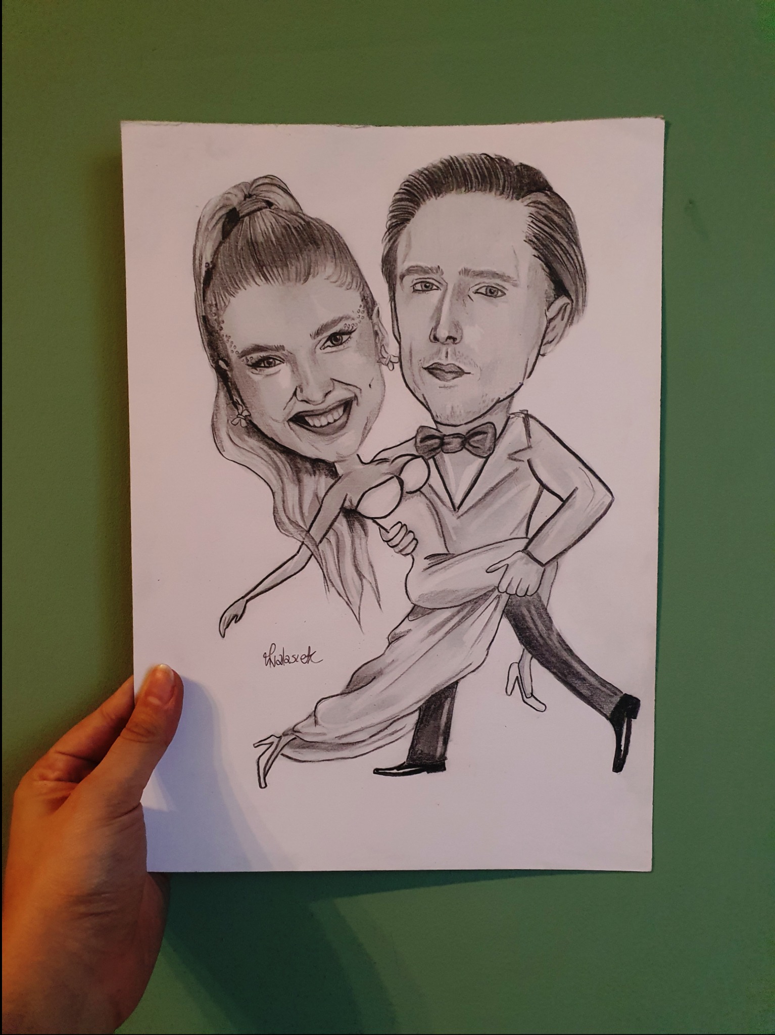 Ręcznie rysowana ołówkiem karykatura dwuosobowa w stylu szkicu, przedstawiająca parę w tańcu tango, umieszczona na białym papierze formatu A4 trzymanym przez rękę na tle zielonej ściany.