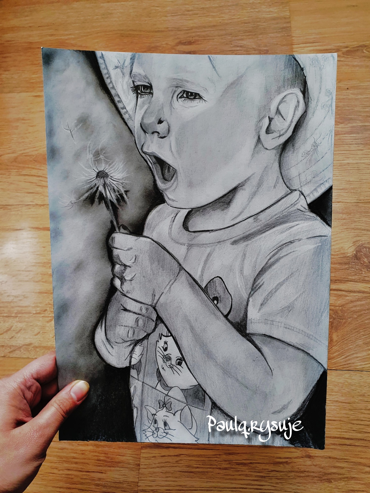 Ręcznie rysowany ołówkiem portret dziecka trzymającego dmuchawca, format A4, widoczny na drewnianym podłożu.