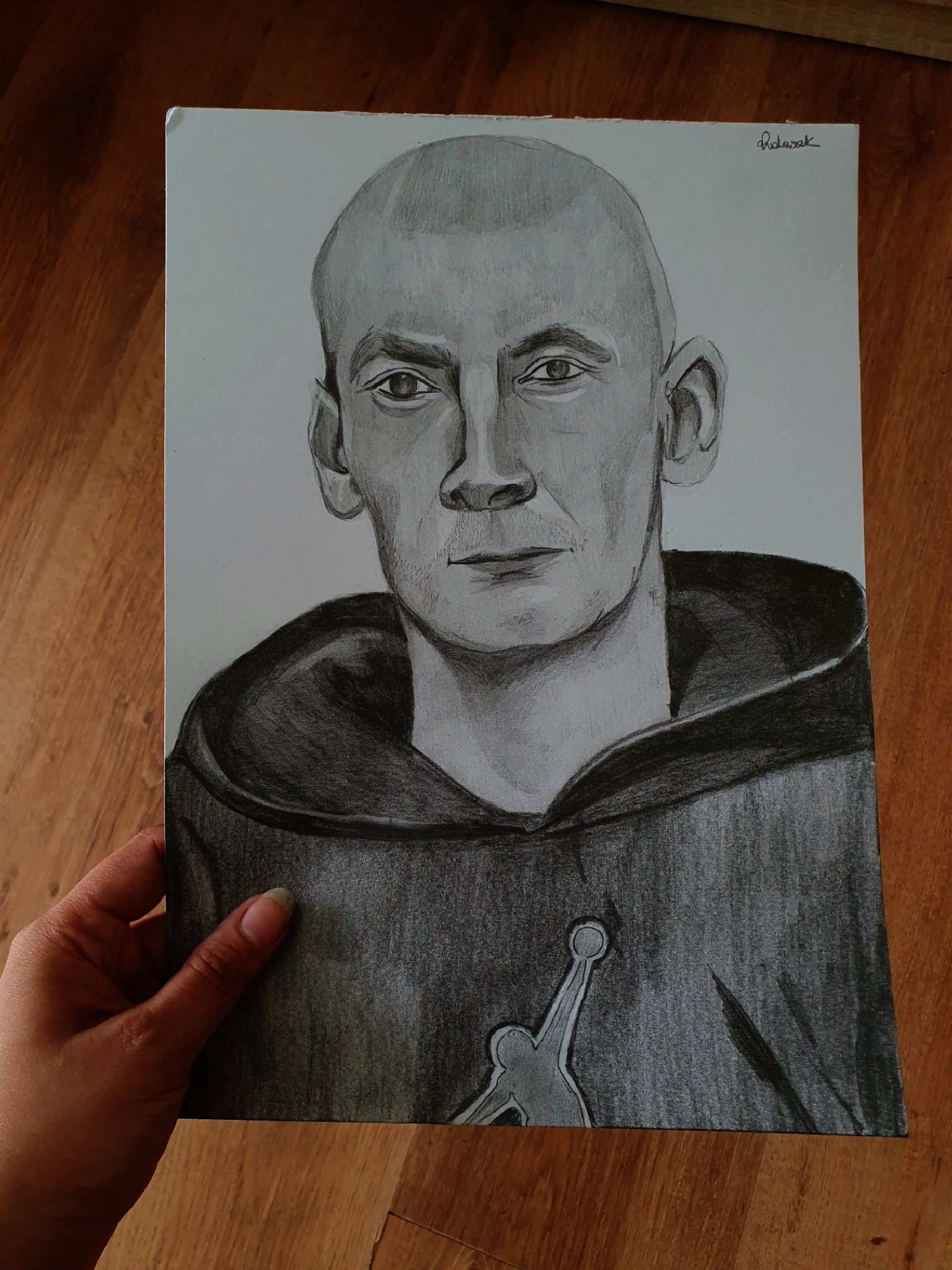 Ręcznie rysowany portret mężczyzny w bluzie z kapturem na formacie A4, trzymany w dłoni na tle drewnianej podłogi.