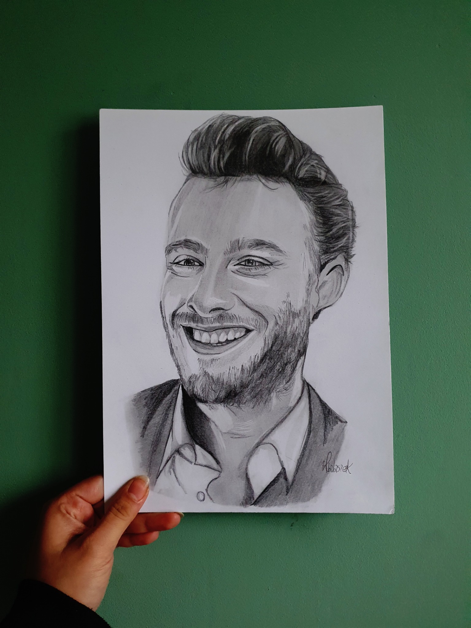 Ręcznie rysowany ołówkiem portret uśmiechniętego mężczyzny w formacie A4, trzymany przed zieloną ścianą, widoczny podpis artysty w prawym dolnym rogu.