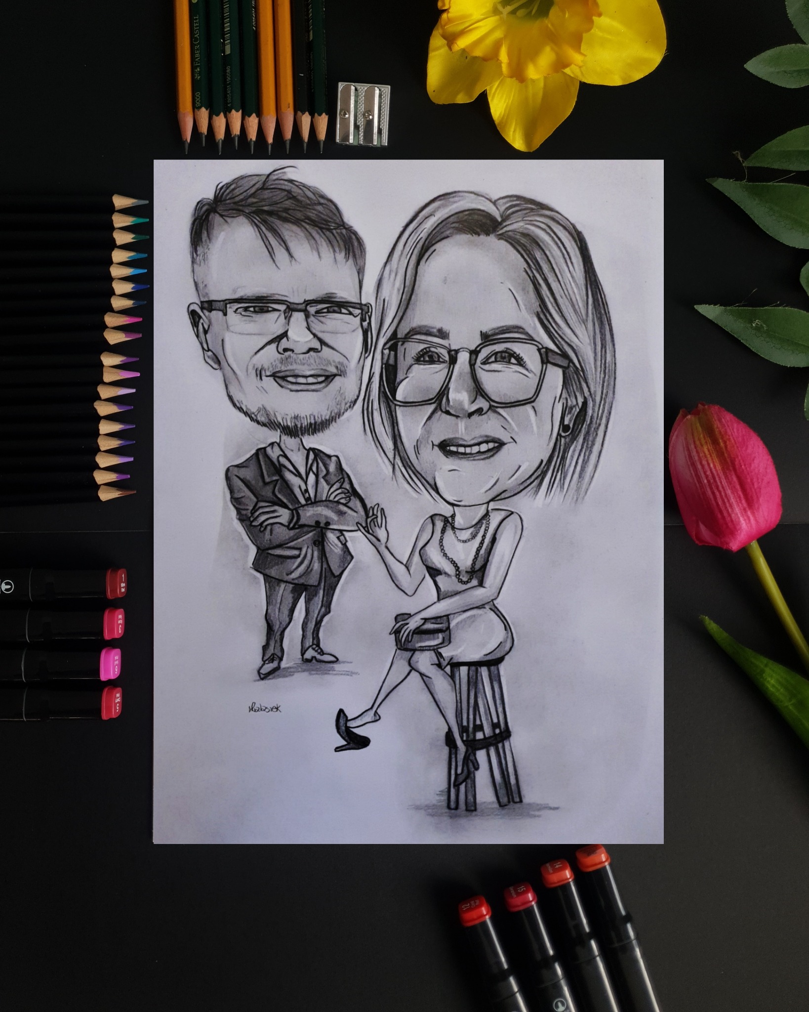 Ręcznie rysowany czarno-biały portret karykaturalny pary na papierze, otoczony kolorowymi kredkami, markerami i kwiatami na czarnym tle.