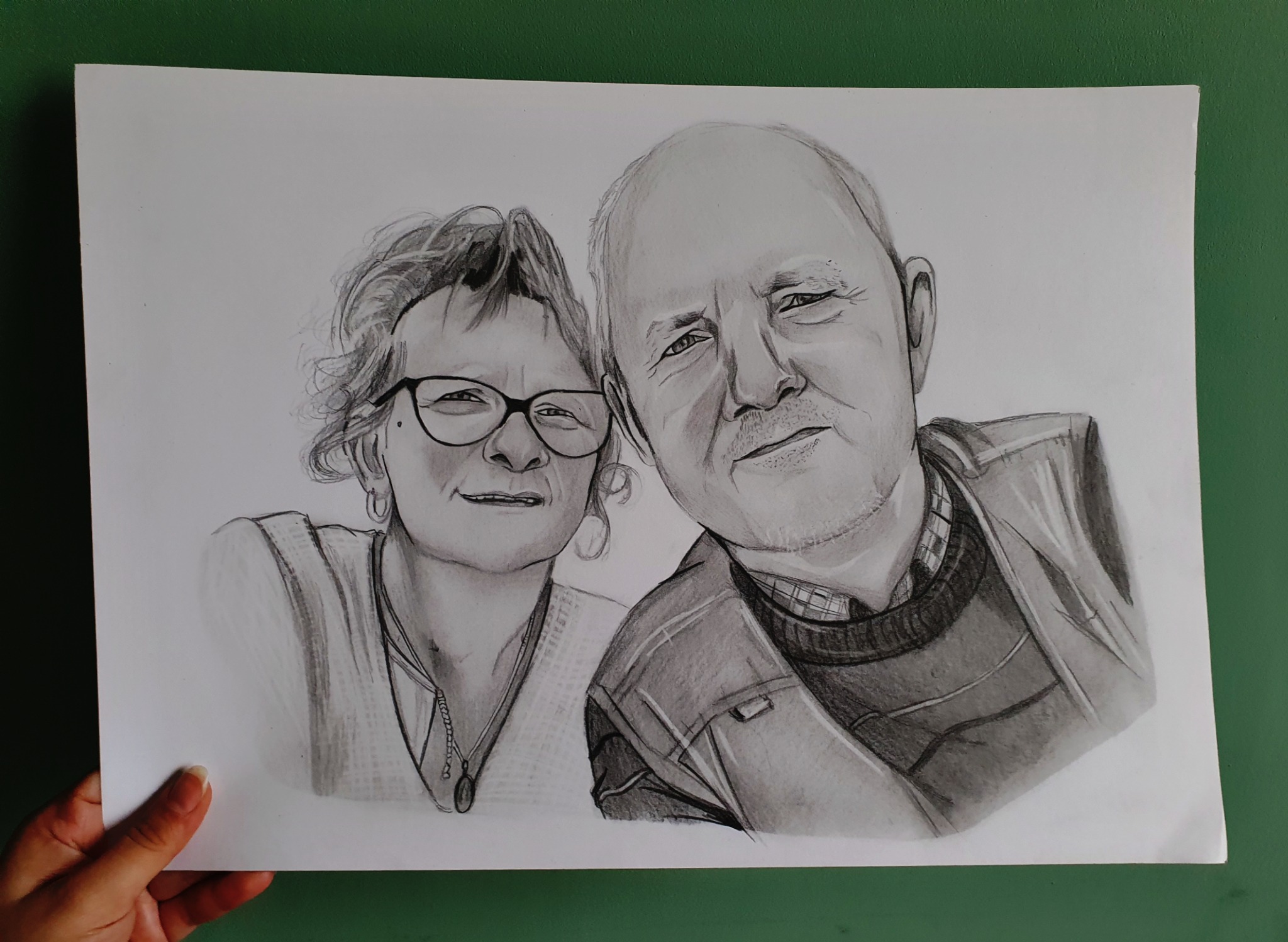 Czarno-biały portret ołówkiem przedstawiający uśmiechniętą parę, trzymany w dłoni na zielonym tle.