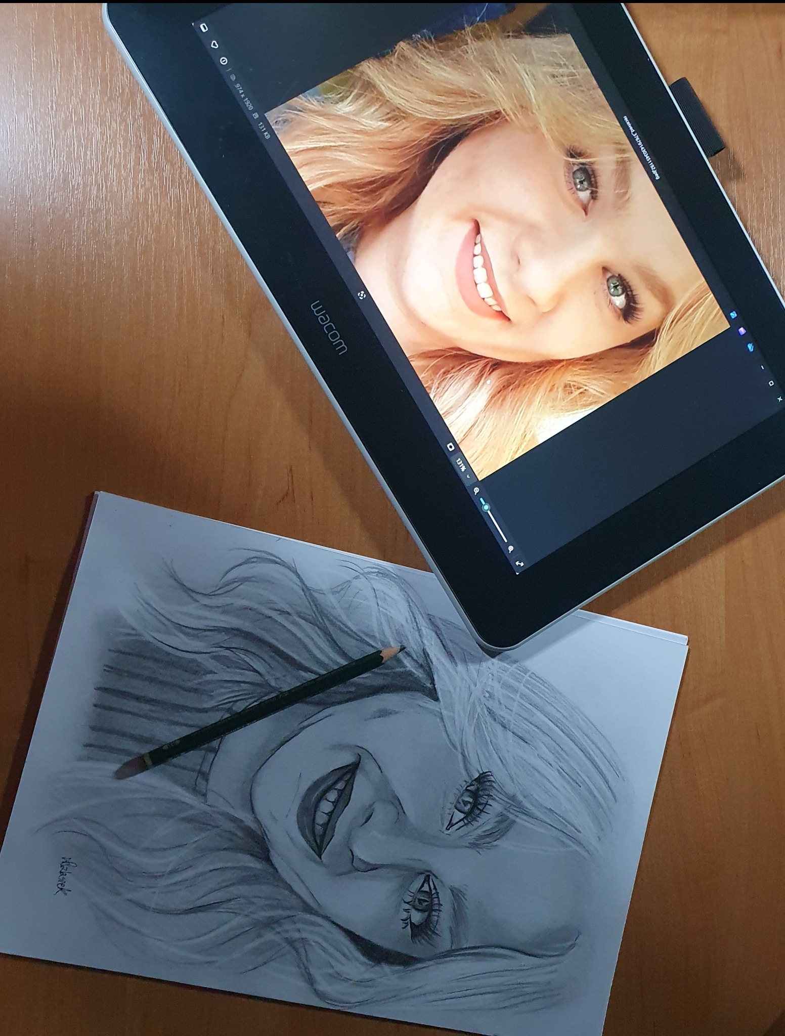 Szkic ołówkiem portretu uśmiechniętej kobiety leżący obok tabletu graficznego Wacom, na którym wyświetlony jest kolorowy portret tej samej osoby.
