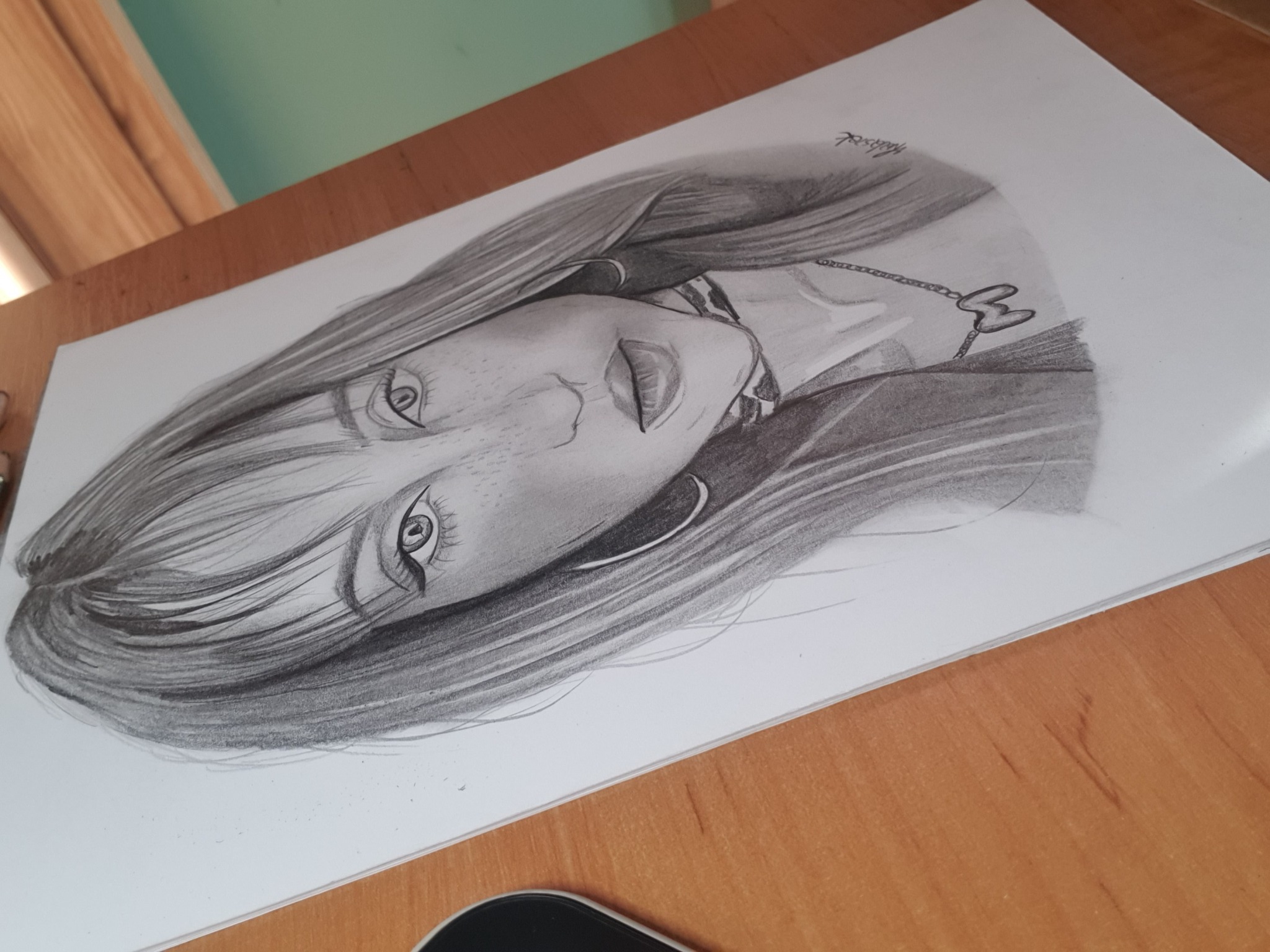 Ołówek na papierze: Portret kobiety z długimi włosami, delikatny cień, widoczne piegi i subtelny naszyjnik z zawieszką w kształcie trójlistnej koniczyny, leżący ukośnie na drewnianym blacie.