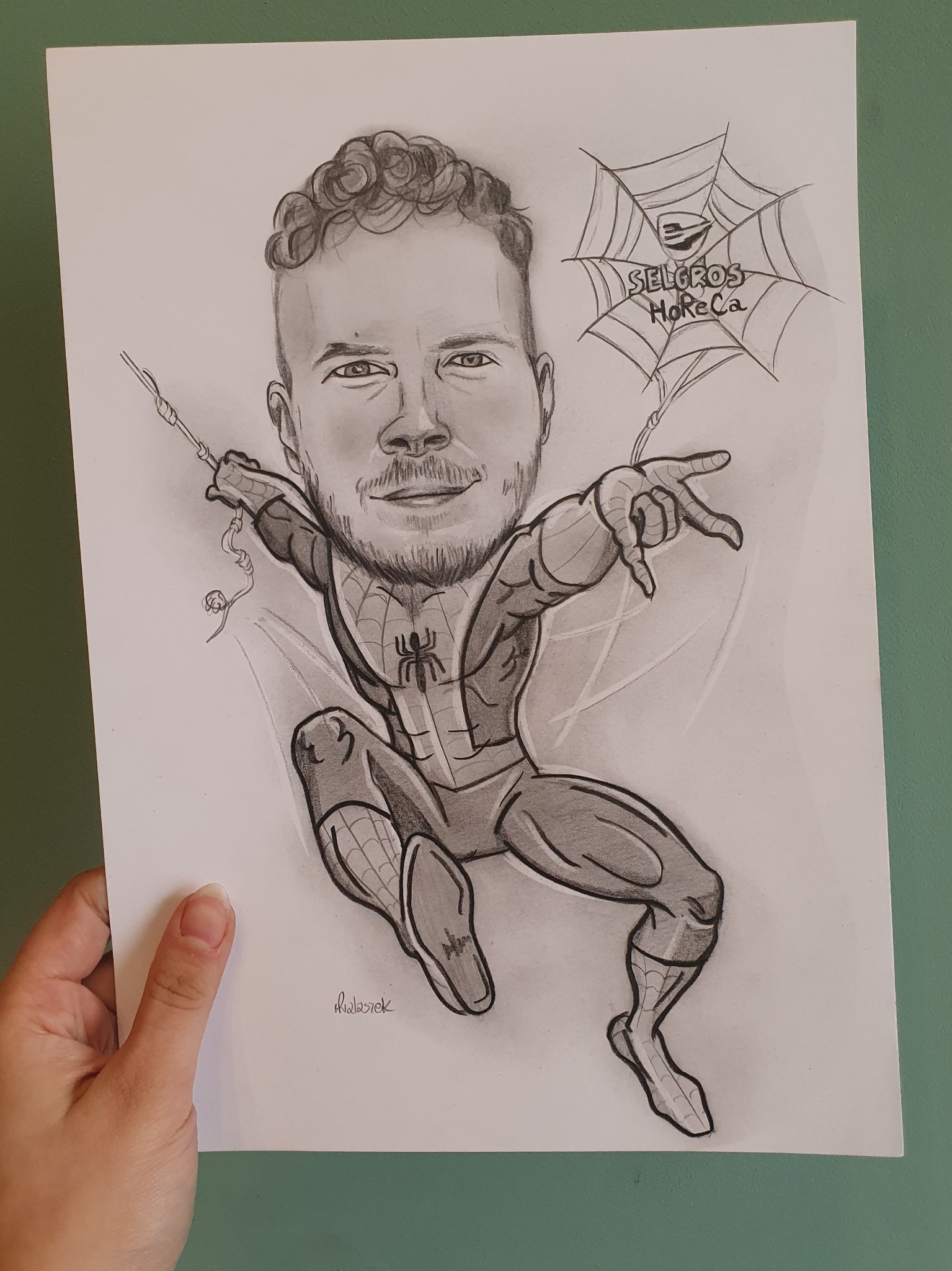 Ręcznie rysowany ołówkiem portret mężczyzny w kostiumie Spider-Mana z elementami logo Selgros HoReCa w pajęczynie, trzymany przez osobę na tle zielonej ściany.