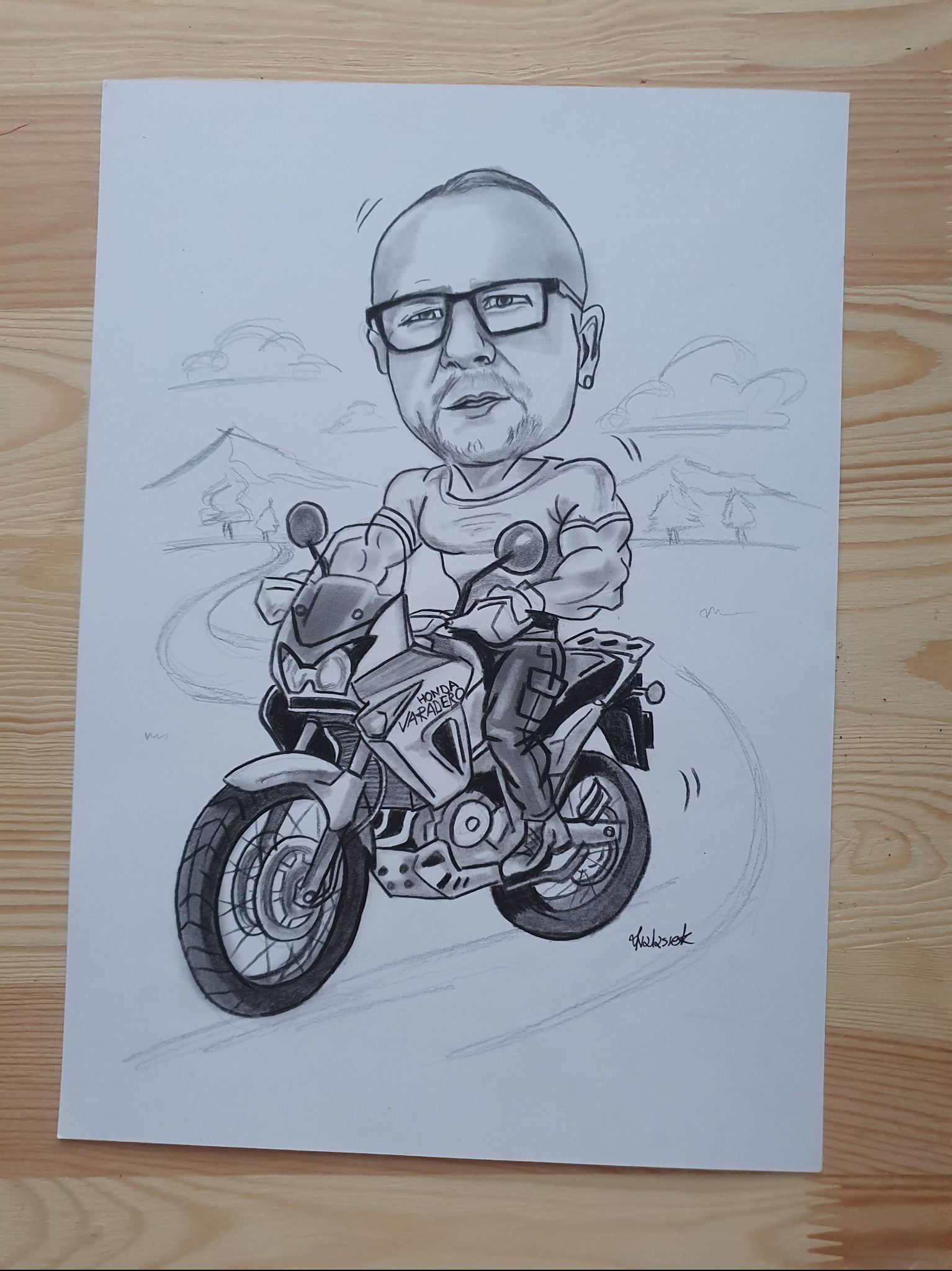Ręcznie rysowany czarno-biały portret karykaturalny mężczyzny na motocyklu Honda Varadero, z tłem górskim i podpisem autora na dole.
