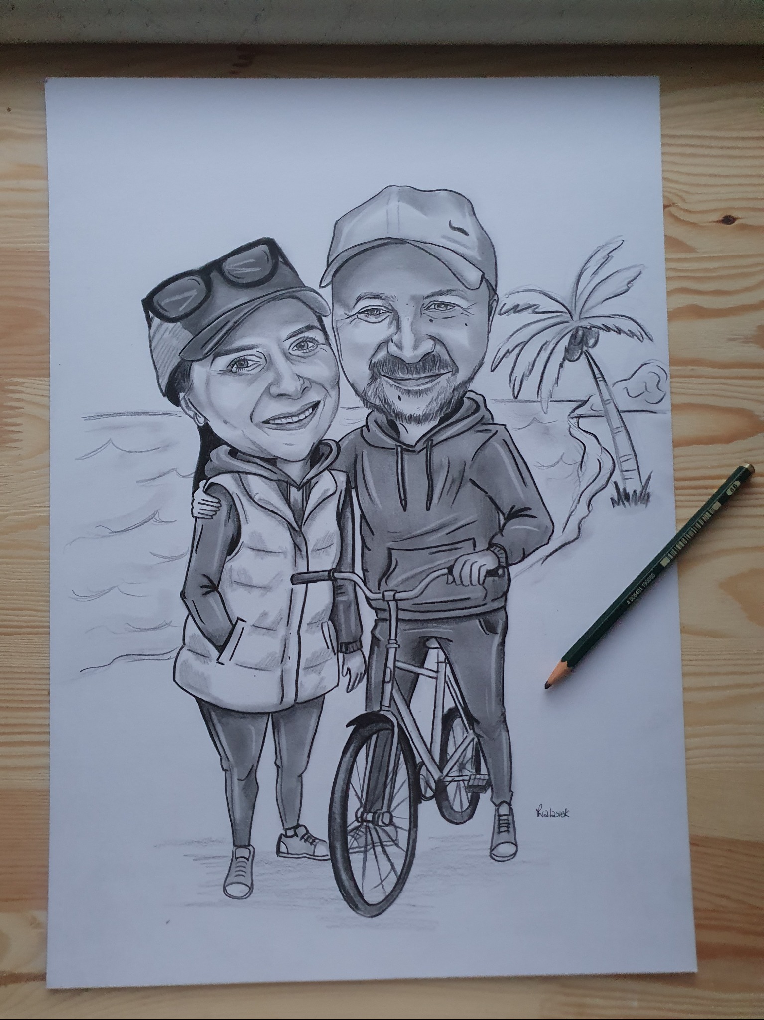Ręcznie rysowany ołówkiem portret pary z rowerem na tle plaży z palmą, leżący na drewnianym stole. Widoczny ołówek obok rysunku. Sygnatura autora w prawym dolnym rogu.