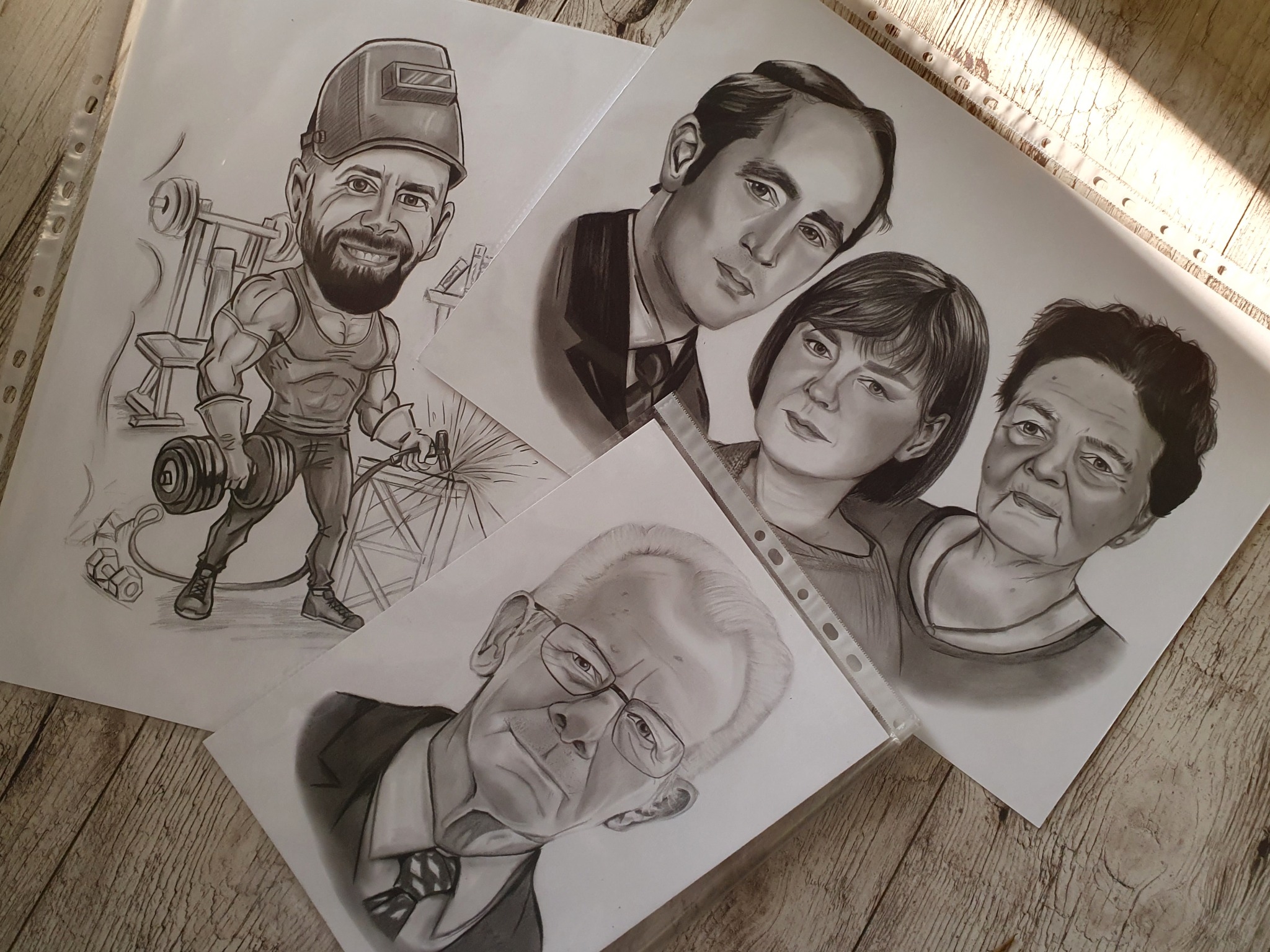 Ręcznie rysowane portrety ołówkiem na białym papierze, przedstawiające karykaturalnego spawacza, portret rodzinny i portret mężczyzny w okularach, leżące na drewnianym podłożu.