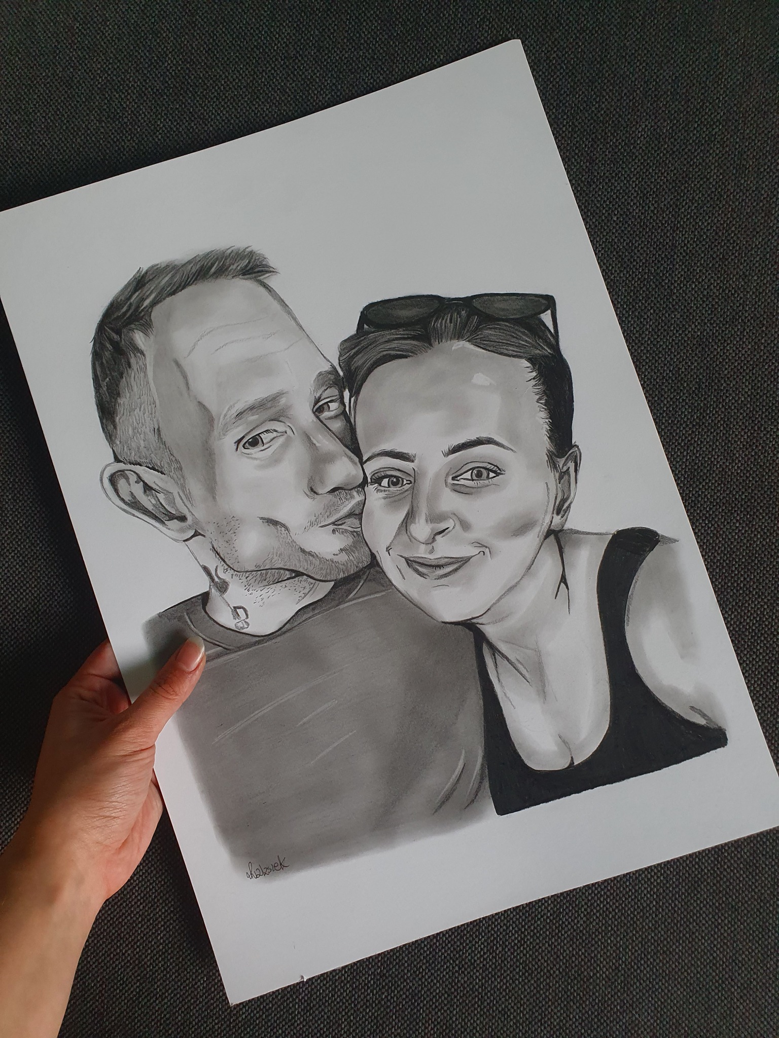 Ręcznie rysowany portret pary w czerni i bieli, trzymany w dłoni. Widoczne detale twarzy, subtelne cieniowanie i sygnatura autora w dolnym rogu.
