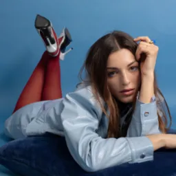 Stylowa sesja portretowa młodej kobiety w błękitnej kurtce, czerwonych rajstopach i szpilkach na poduszce, na jednolitym niebieskim tle. Uwagę przykuwają detale makijażu i biżuterii.