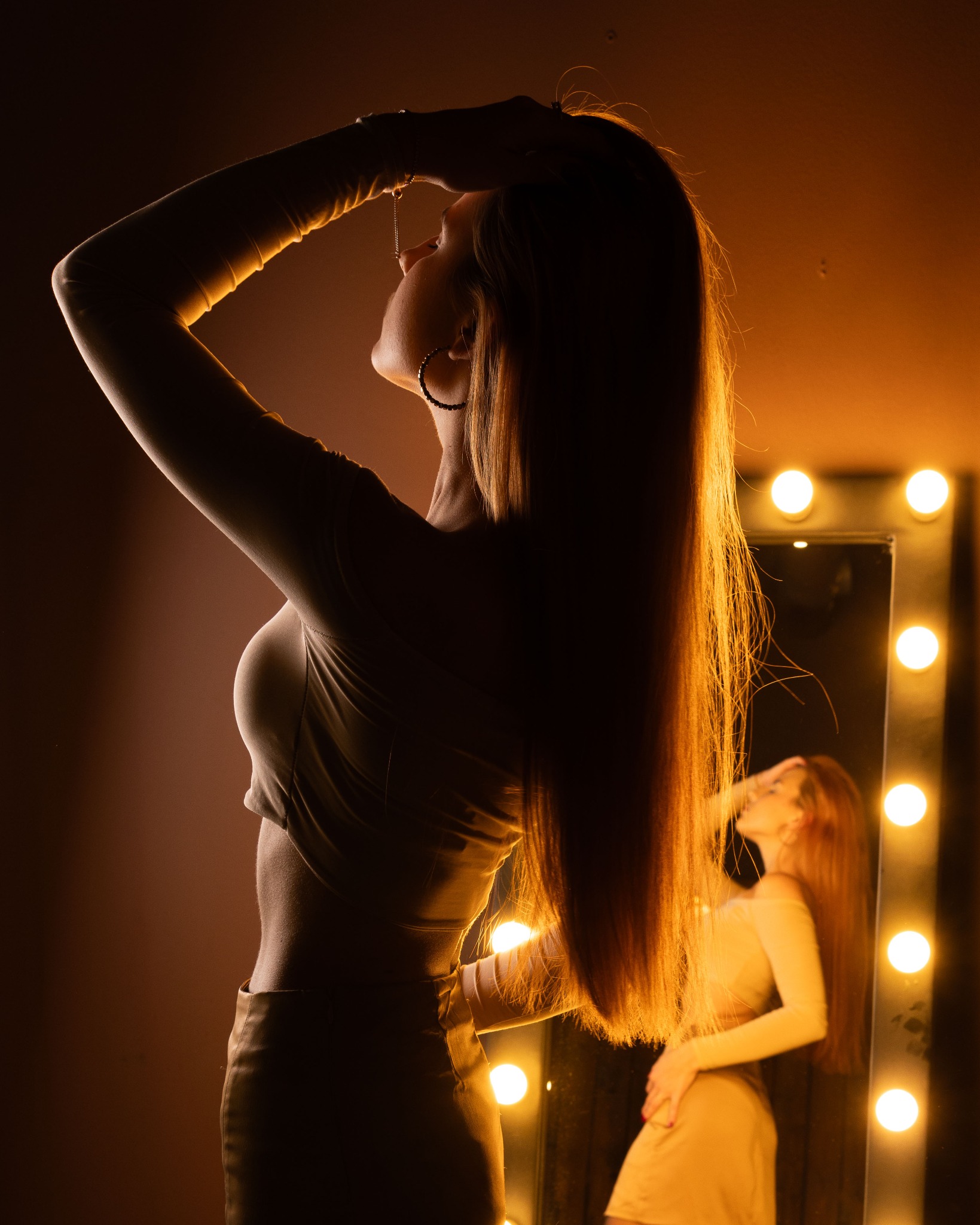 Kobieta w obcisłym stroju pozuje przed lustrem z żarówkami, oświetlona ciepłym światłem, z widocznym profilem i długimi, prostymi włosami.