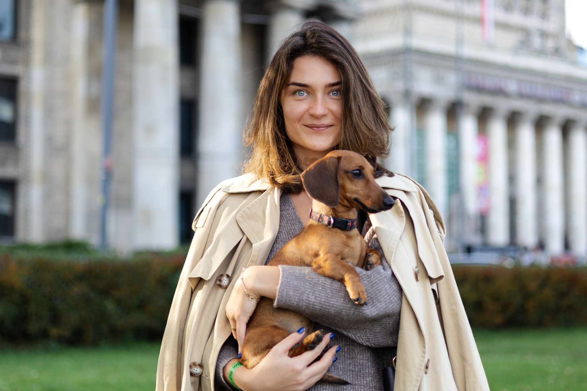 Uśmiechnięta kobieta w beżowym płaszczu trzyma brązowego jamnika na tle budynku z kolumnami.