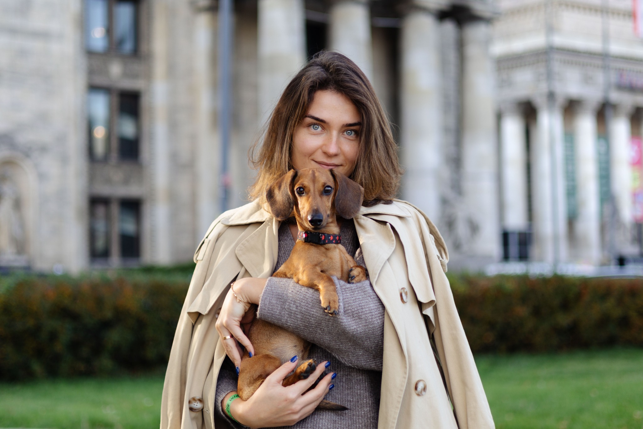 Portret kobiety w beżowym płaszczu trzymającej jamnika na tle budynku z kolumnami