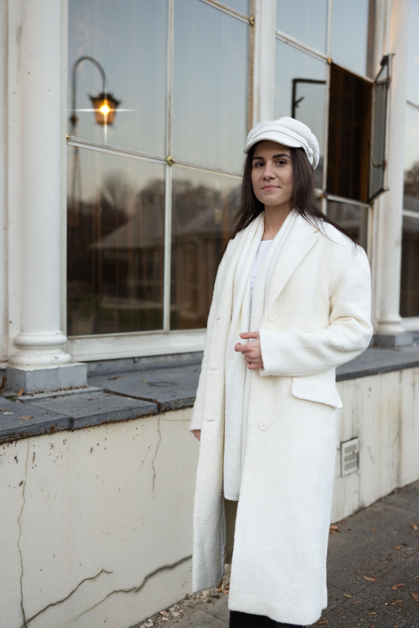 Kobieta w białym płaszczu i czapce pozuje na tle budynku z dużymi oknami, w których odbija się latarnia.