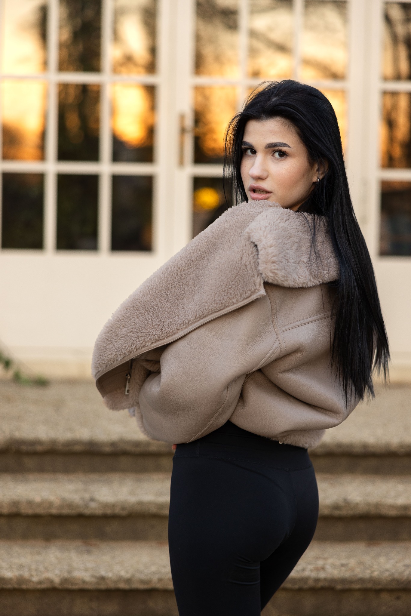 Kobieta z długimi czarnymi włosami, ubrana w beżową kurtkę typu shearling i czarne legginsy, odwraca się przez ramię na tle białych drzwi i kamiennych schodów.