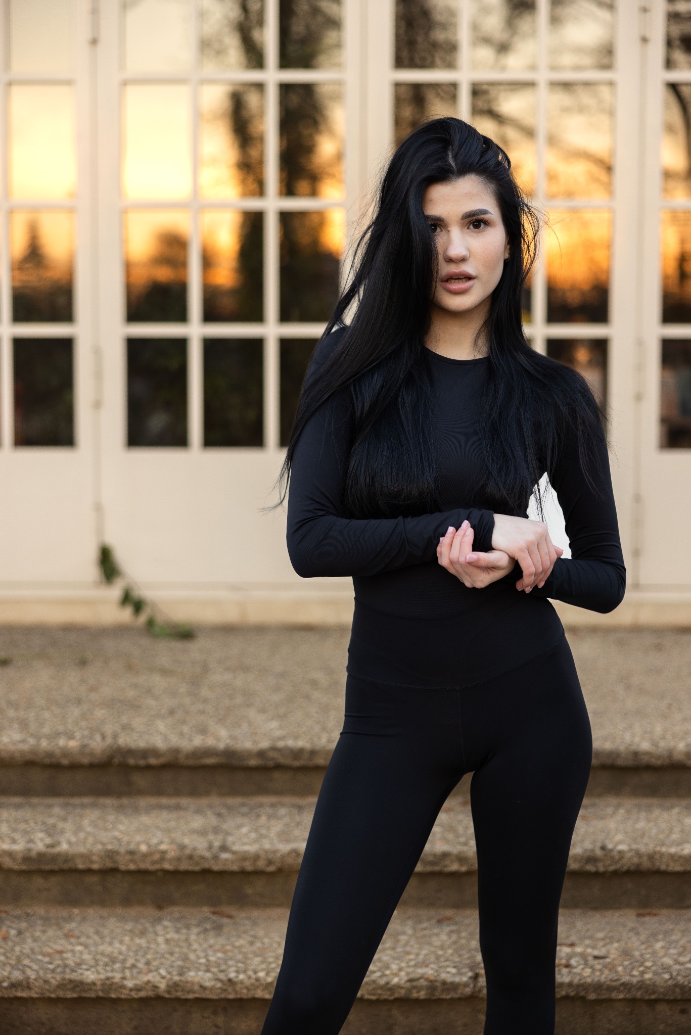 Portret kobiety w czarnym stroju sportowym na tle białych drzwi z szybami, z widocznym zachodem słońca.