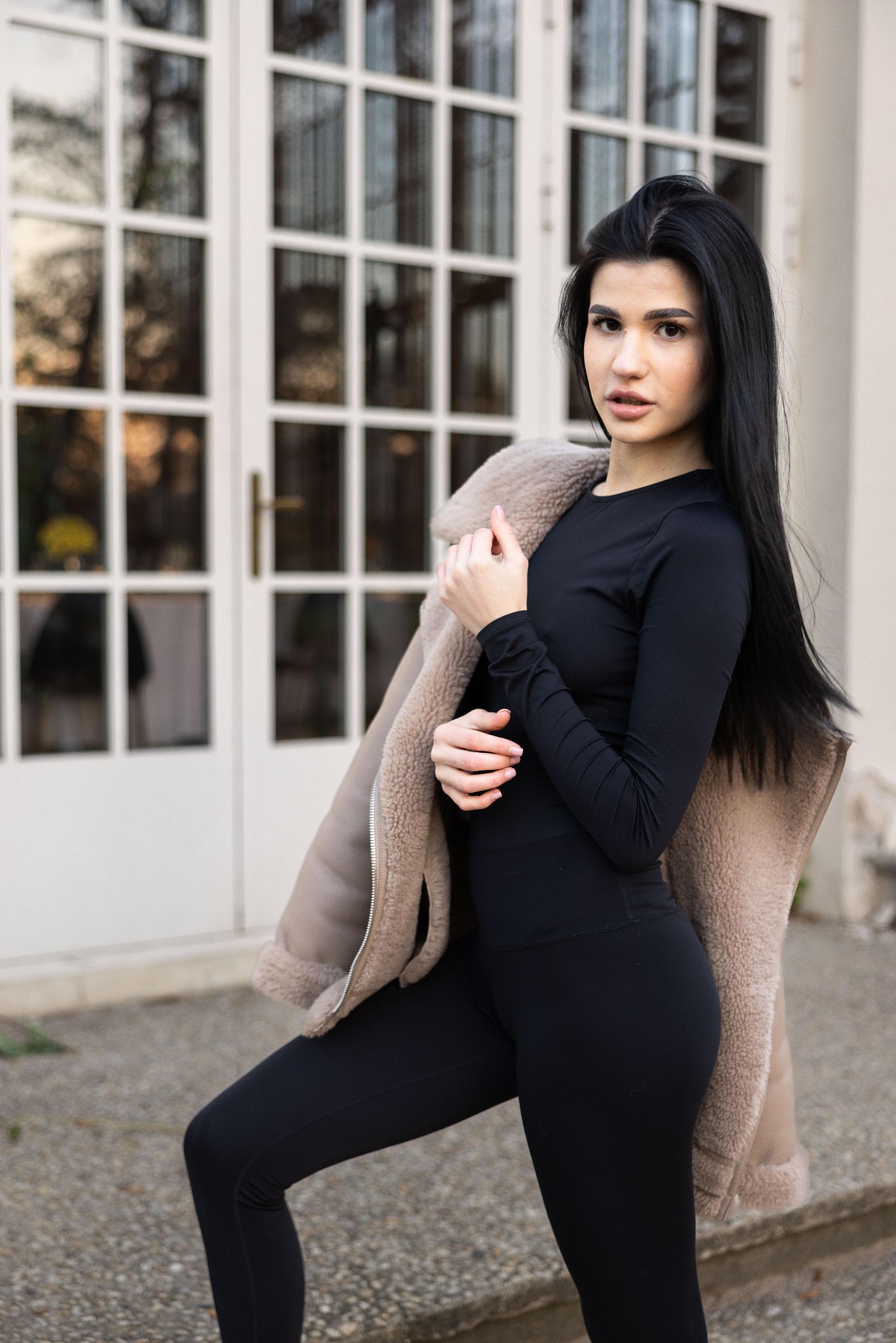 Portret młodej kobiety w czarnym stroju sportowym, z kurtką z kożuszkiem na ramieniu, pozującej na tle białych drzwi z licznymi szybkami.