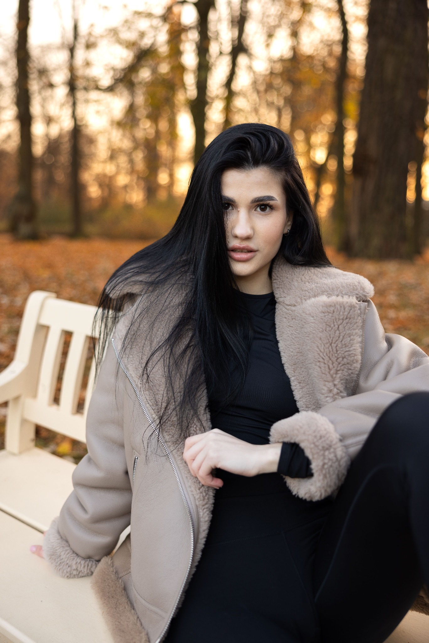 Portret kobiety o długich, czarnych włosach w beżowej kurtce z futrzanym kołnierzem, siedzącej na białej ławce w jesiennym parku, z drzewami i opadłymi liśćmi w tle.