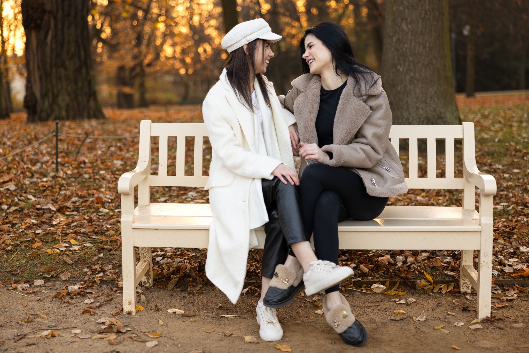Dwie uśmiechnięte kobiety siedzą na białej ławce w parku jesienią, w tle drzewa z opadającymi liśćmi, oświetlone popołudniowym słońcem.
