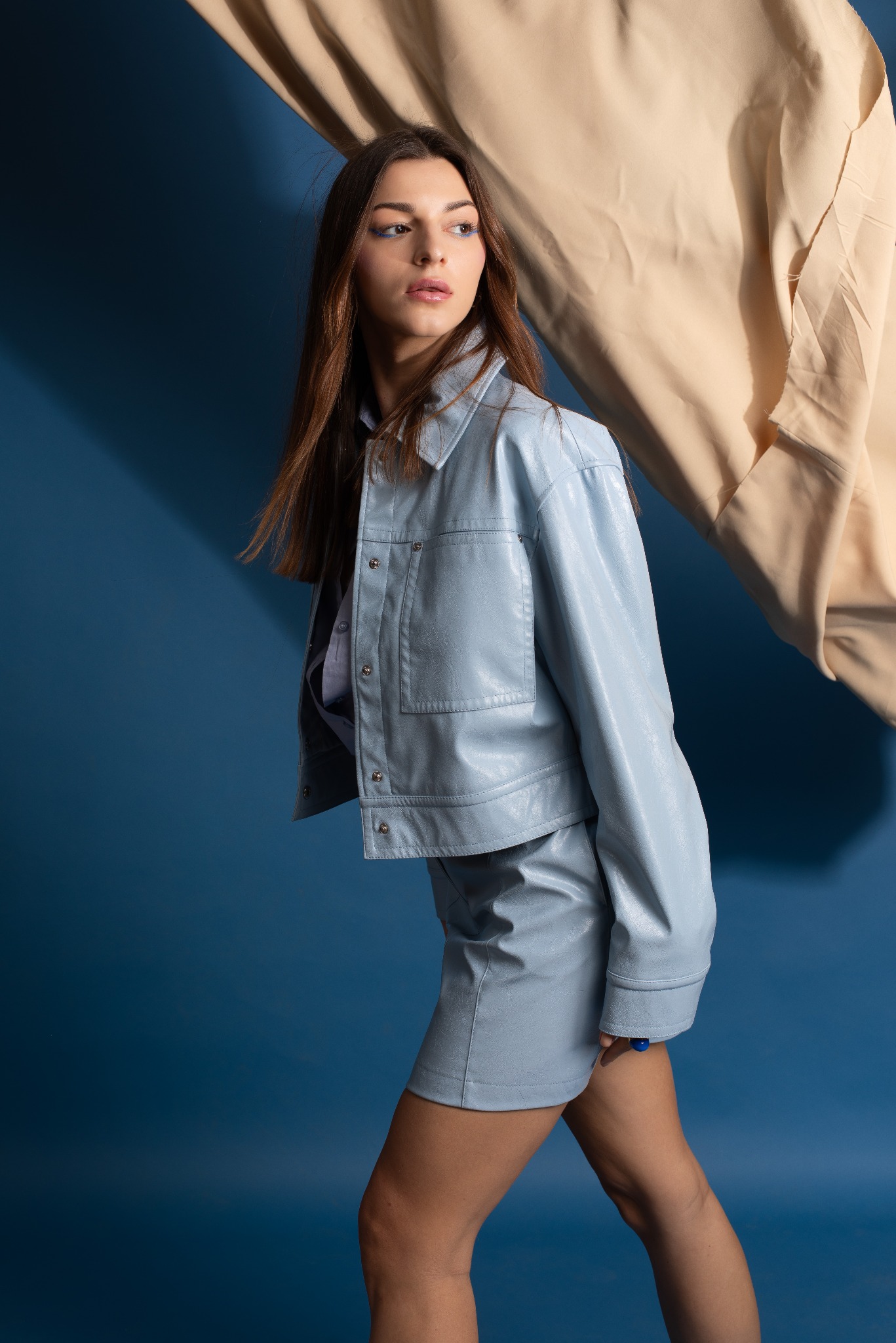 Kobieta w błękitnym skórzanym komplecie (krótka kurtka i spódnica) na tle błękitnej ściany i beżowej tkaniny, z wyrazistym niebieskim makijażem oczu, patrzy w bok.