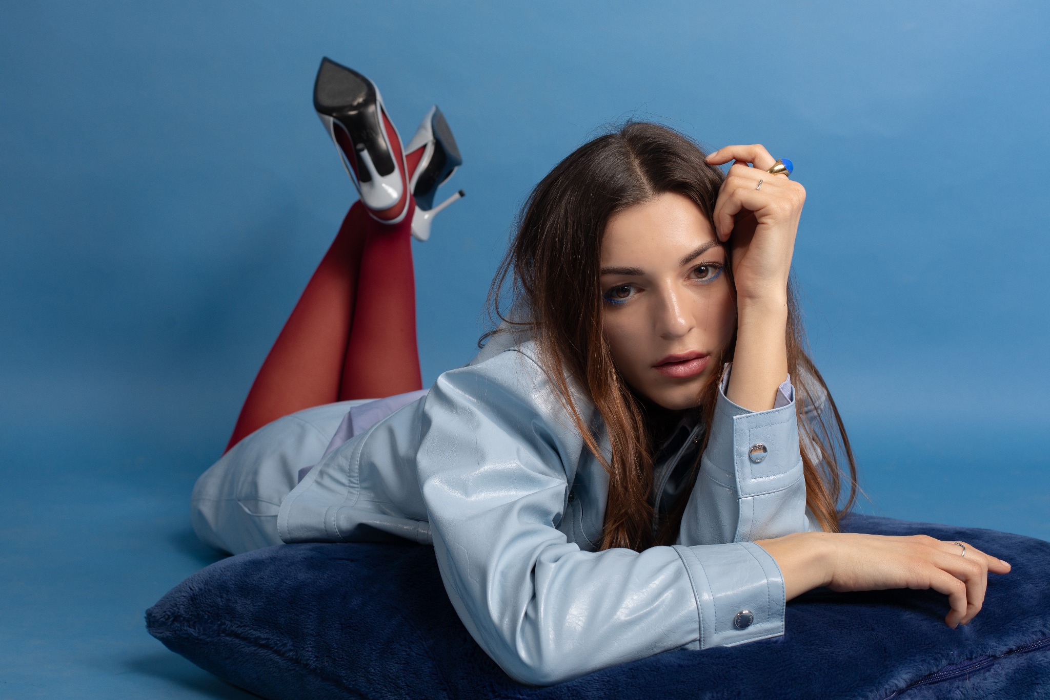Stylowa sesja portretowa młodej kobiety w błękitnej kurtce, czerwonych rajstopach i szpilkach na poduszce, na jednolitym niebieskim tle. Uwagę przykuwają detale makijażu i biżuterii.