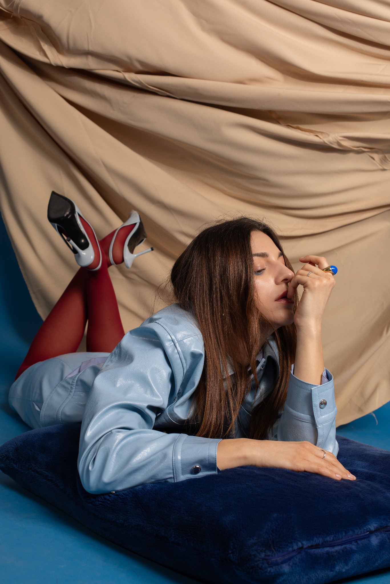 Kobieta w błękitnej kurtce leży na poduszce, czerwone rajstopy i białe szpilki. Beżowe tło. Ujęcie z profilu, dłoń przy twarzy, pierścionek z niebieskim kamieniem.