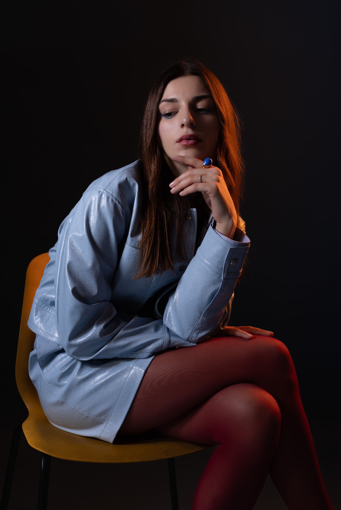 Portret kobiety siedzącej na żółtym krześle, ubrana w błękitny skórzany komplet, czerwone rajstopy, z niebieskim pierścieniem na palcu, na ciemnym tle.