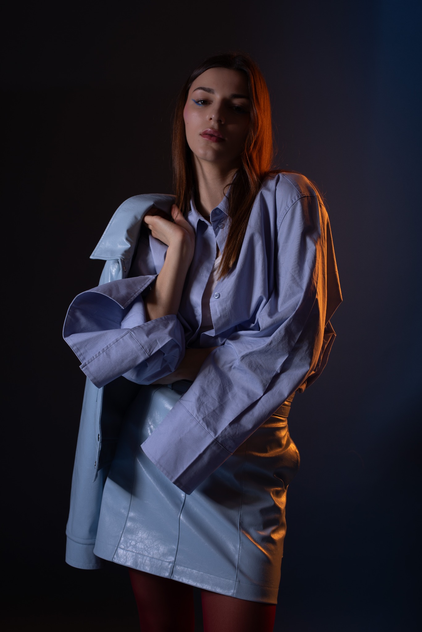 Portret kobiety w niebieskiej, skórzanej spódnicy i koszuli, z marynarką zarzuconą na ramię, pozującej na ciemnym tle z pomarańczowo-niebieskim oświetleniem.