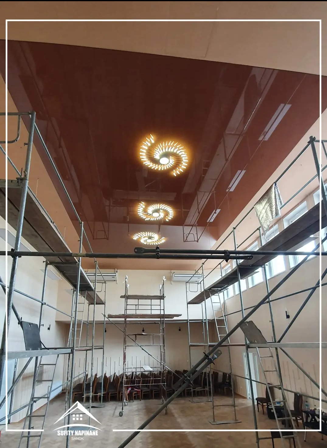 Brązowy, błyszczący sufit z trzema spiralnymi lampami widziany spod rusztowania w trakcie montażu.