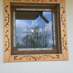 Zubek Frez CNC - Okno z drewnianą ramą zdobioną motywami roślinnymi, widok na ogród przez szybę.