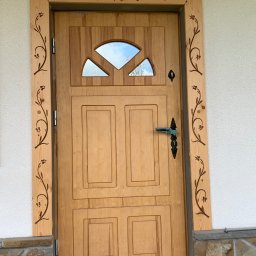 Zubek Frez CNC - Drewniane drzwi zewnętrzne z ozdobną ramą i szybą w górnej części, wyposażone w czarną klamkę i zamek, zamontowane w białej ścianie z kamienną podmurówką.