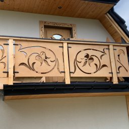 Zubek Frez CNC - Balustrada balkonowa z jasnego drewna z wyciętymi motywami roślinnymi, czarne okucia zawiasowe, w tle fragment elewacji budynku z drewnianą stolarką okienną z motywami kwiatowymi.