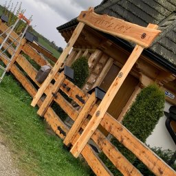Zubek Frez CNC - Nowe, jasne ogrodzenie z drewna w stylu góralskim z ozdobnymi elementami i napisem 'WITAM' nad wejściem, częściowo zasłaniające budynek, z drabinką opartą o bok ogrodzenia.