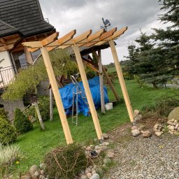 Zubek Frez CNC - Konstrukcja drewniana altany w trakcie budowy, z widoczną niebieską plandeką, drabiną i dekoracyjnym wiatrowskazem w kształcie koguta na dachu, na tle domu z ciemnym dachem i zielonego ogrodu.