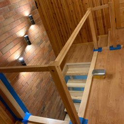 Zubek Frez CNC - Wnętrze z drewnianymi schodami w trakcie montażu, zabezpieczone niebieską taśmą malarską, widoczna puszka z preparatem do drewna, ściana z cegły i drewniana boazeria.