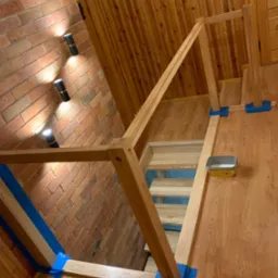 Wnętrze z drewnianymi schodami w trakcie montażu, zabezpieczone niebieską taśmą malarską, widoczna puszka z preparatem do drewna, ściana z cegły i drewniana boazeria.