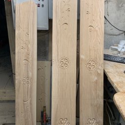 Zubek Frez CNC - Trzy drewniane panele z wyżłobionymi wzorami kwiatowymi i geometrycznymi, ustawione pionowo w warsztacie, z widocznym panelem sterowania maszyny w tle.